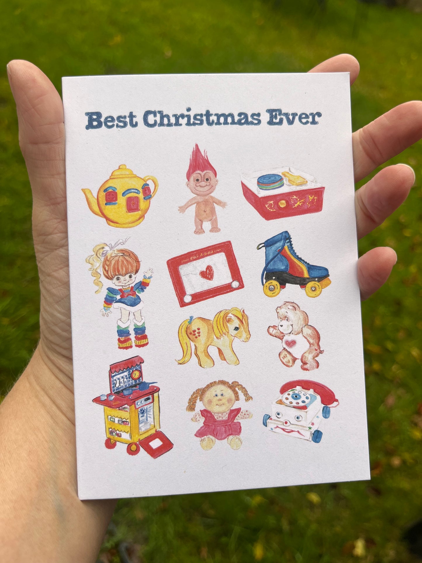 Hand holds Best Christmas Ever cards featuring retro toy illustrations including red haired troll doll, etcher sketch and roller skates