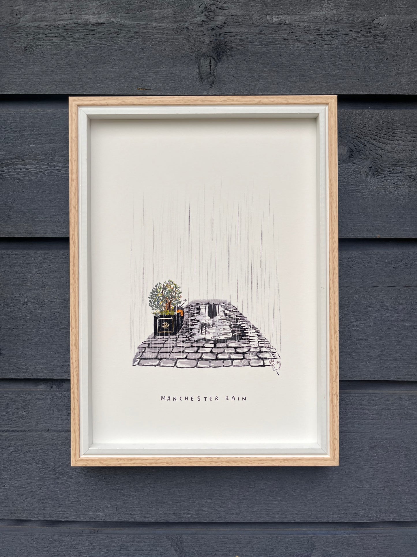 Manchester rain art print framed in a light wooden frame hung on a wooden dark grey panelled wall