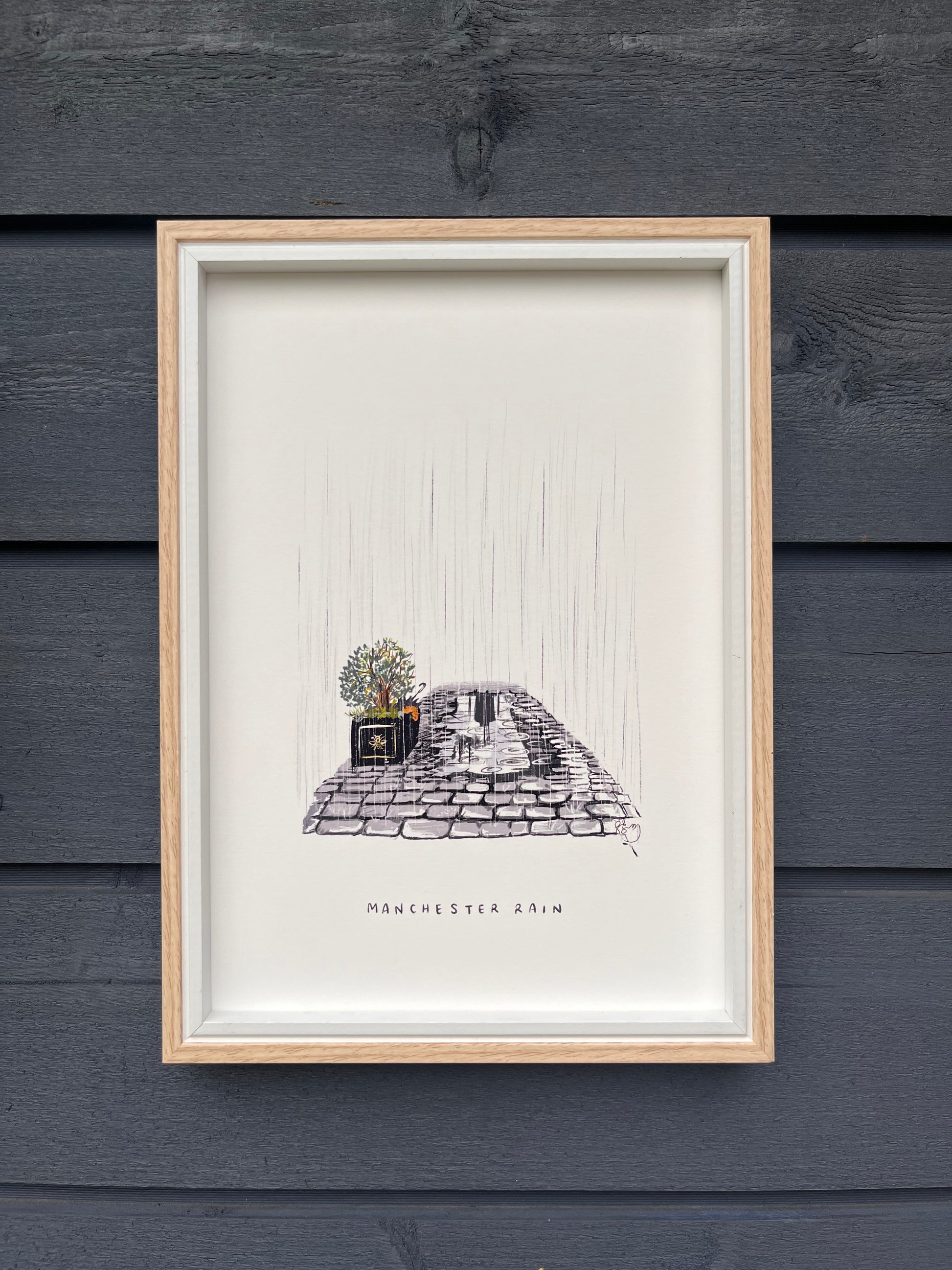 Manchester rain art print framed in a light wooden frame hung on a wooden dark grey panelled wall