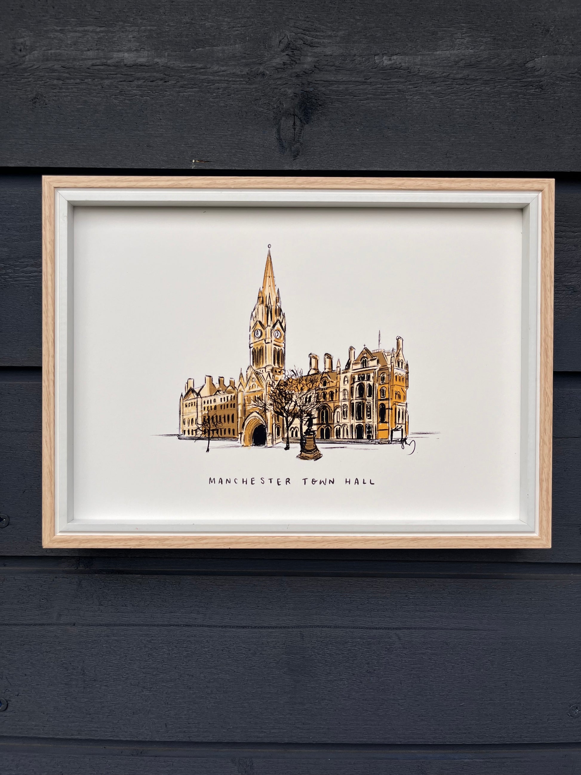 Manchester town hall illustrated art print, framed in a light wooden frame hung on a wooden dark grey panelled wall